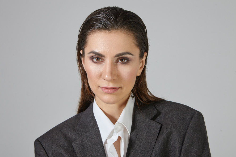 Kobieta w eleganckim szarym garniturze i białej koszuli, z profesjonalnym wyrazem twarzy, na neutralnym tle.