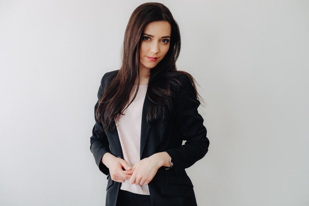 Młoda kobieta w eleganckim czarnym garniturze i jasnej bluzce, stojąca na jasnym tle z delikatnym uśmiechem.