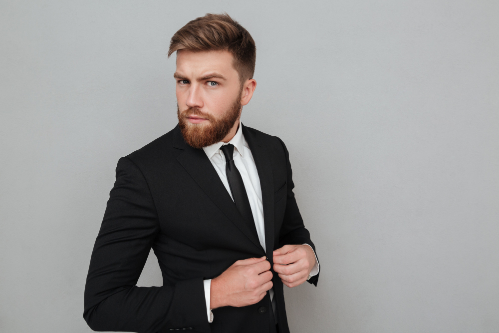 Mężczyzna z brodą w eleganckim czarnym garniturze i krawacie, poprawiający marynarkę, na jasnym tle.