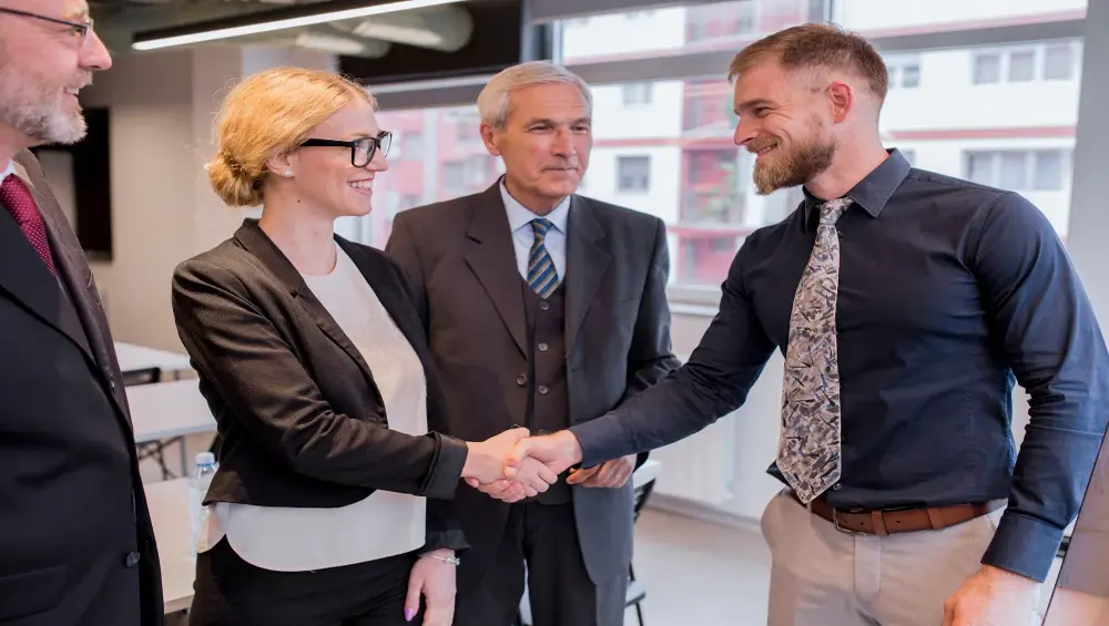 Grupa czterech osób w formalnych strojach w biurze. Kobieta w okularach i mężczyzna z brodą uśmiechają się i podają sobie ręce. W tle dwóch starszych mężczyzn w garniturach obserwuje sytuację.